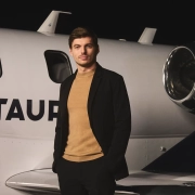 Max Verstappen stoji ob zasebnem letalu z napisom AlphaTauri, elegantno oblečen v črno jakno in bež pulover, kot novi globalni ambasador modne znamke AlphaTauri. / Max Verstappen standing next to a private jet with the AlphaTauri logo, dressed in a black jacket and beige sweater, as the new global ambassador for AlphaTauri fashion brand.