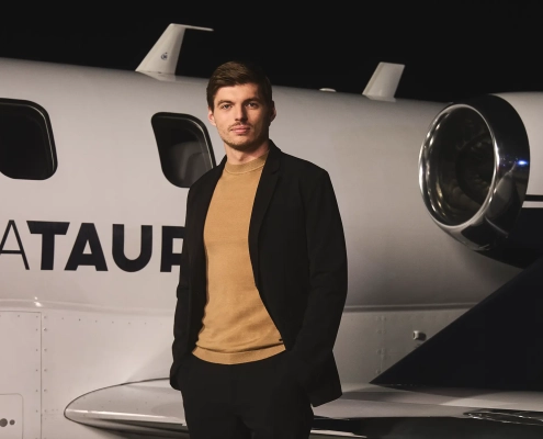 Max Verstappen stoji ob zasebnem letalu z napisom AlphaTauri, elegantno oblečen v črno jakno in bež pulover, kot novi globalni ambasador modne znamke AlphaTauri. / Max Verstappen standing next to a private jet with the AlphaTauri logo, dressed in a black jacket and beige sweater, as the new global ambassador for AlphaTauri fashion brand.