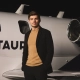 Max Verstappen stoji ob zasebnem letalu z napisom AlphaTauri, elegantno oblečen v črno jakno in bež pulover, kot novi globalni ambasador modne znamke AlphaTauri. / Max Verstappen standing next to a private jet with the AlphaTauri logo, dressed in a black jacket and beige sweater, as the new global ambassador for AlphaTauri fashion brand.