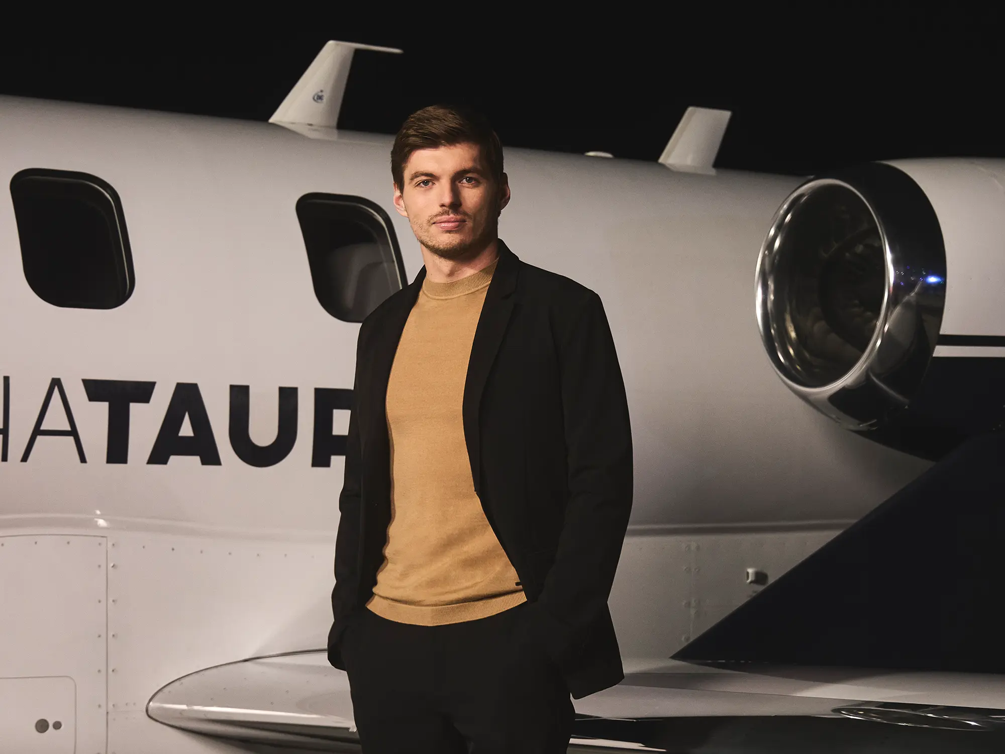 Max Verstappen stoji ob zasebnem letalu z napisom AlphaTauri, elegantno oblečen v črno jakno in bež pulover, kot novi globalni ambasador modne znamke AlphaTauri. / Max Verstappen standing next to a private jet with the AlphaTauri logo, dressed in a black jacket and beige sweater, as the new global ambassador for AlphaTauri fashion brand.