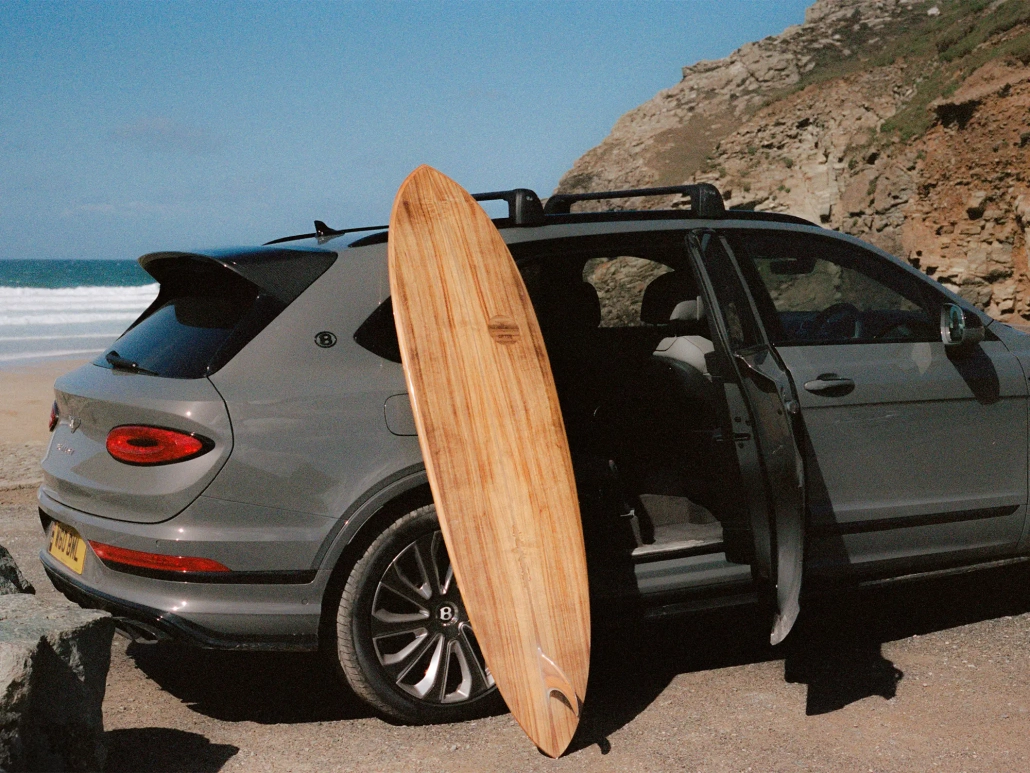 Ročno izdelana deska za deskanje iz Bentleyjevega furnirja Koa, naslonjena na luksuzni SUV Bentley Bentayga, parkiran ob obali s pogledom na valove. / Handcrafted surfboard made from Bentley’s Koa veneer, leaning against a luxury Bentley Bentayga SUV parked by the coast with waves in the background.