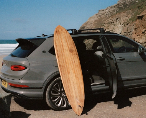 Ročno izdelana deska za deskanje iz Bentleyjevega furnirja Koa, naslonjena na luksuzni SUV Bentley Bentayga, parkiran ob obali s pogledom na valove. / Handcrafted surfboard made from Bentley’s Koa veneer, leaning against a luxury Bentley Bentayga SUV parked by the coast with waves in the background.