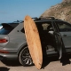 Ročno izdelana deska za deskanje iz Bentleyjevega furnirja Koa, naslonjena na luksuzni SUV Bentley Bentayga, parkiran ob obali s pogledom na valove. / Handcrafted surfboard made from Bentley’s Koa veneer, leaning against a luxury Bentley Bentayga SUV parked by the coast with waves in the background.