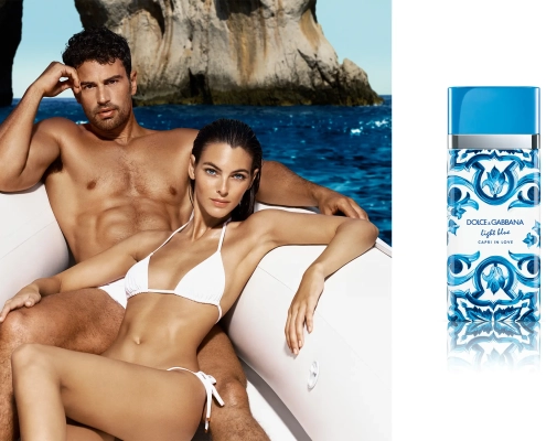 Igralec Theo James in manekenka v beli kopalkah na gumenjaku pred obalo Caprija, ob njima steklenička parfuma Dolce & Gabbana Light Blue Capri in Love. / Actor Theo James and a model in white swimwear on an inflatable boat near Capri, with a Dolce & Gabbana Light Blue Capri in Love perfume bottle beside them.