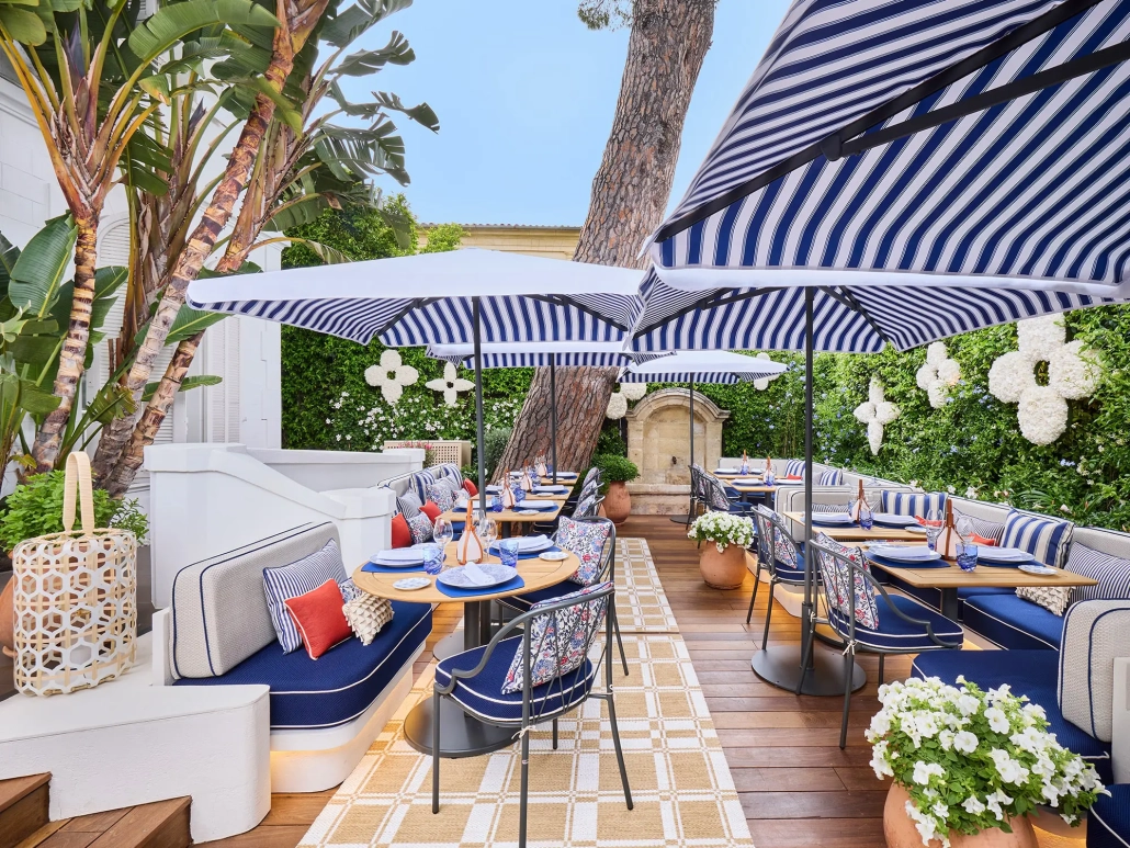 Elegantna zunanja restavracija Louis Vuitton v St. Tropezu z modro-belimi črtastimi senčniki, cvetličnimi aranžmaji in prefinjenim sedežnim pohištvom v sredozemskem slogu. / Elegant outdoor Louis Vuitton restaurant in St. Tropez featuring blue-and-white striped umbrellas, floral decor, and refined Mediterranean-style seating.