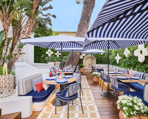 Elegantna zunanja restavracija Louis Vuitton v St. Tropezu z modro-belimi črtastimi senčniki, cvetličnimi aranžmaji in prefinjenim sedežnim pohištvom v sredozemskem slogu. / Elegant outdoor Louis Vuitton restaurant in St. Tropez featuring blue-and-white striped umbrellas, floral decor, and refined Mediterranean-style seating.