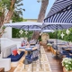 Elegantna zunanja restavracija Louis Vuitton v St. Tropezu z modro-belimi črtastimi senčniki, cvetličnimi aranžmaji in prefinjenim sedežnim pohištvom v sredozemskem slogu. / Elegant outdoor Louis Vuitton restaurant in St. Tropez featuring blue-and-white striped umbrellas, floral decor, and refined Mediterranean-style seating.