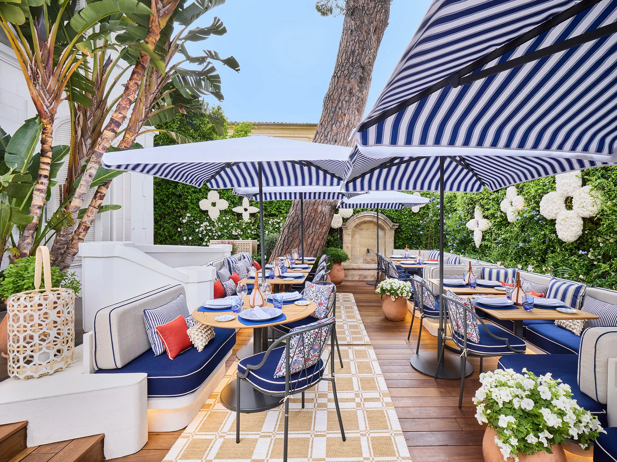 Elegantna zunanja restavracija Louis Vuitton v St. Tropezu z modro-belimi črtastimi senčniki, cvetličnimi aranžmaji in prefinjenim sedežnim pohištvom v sredozemskem slogu. / Elegant outdoor Louis Vuitton restaurant in St. Tropez featuring blue-and-white striped umbrellas, floral decor, and refined Mediterranean-style seating.