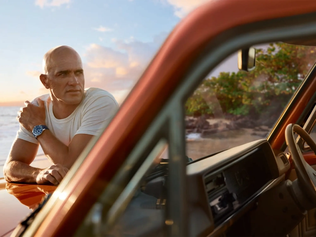 Kelly Slater ob obali med sončnim zahodom, naslonjen na retro terensko vozilo, nosi uro Breitling Superocean Heritage B31 Automatic 40 Kelly Slater Limited Edition. / Kelly Slater leaning on a retro truck at sunset, wearing the Breitling Superocean Heritage B31 Automatic 40 Kelly Slater Limited Edition watch.