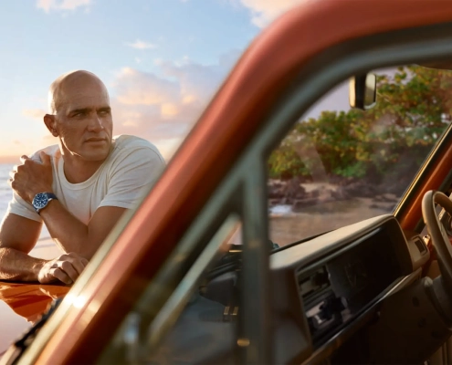 Kelly Slater ob obali med sončnim zahodom, naslonjen na retro terensko vozilo, nosi uro Breitling Superocean Heritage B31 Automatic 40 Kelly Slater Limited Edition. / Kelly Slater leaning on a retro truck at sunset, wearing the Breitling Superocean Heritage B31 Automatic 40 Kelly Slater Limited Edition watch.