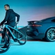 Moški ob Porsche eBike Cross Performance EXC v barvi Provence poleg modrega Porsche Taycana Turbo GT v umetniško osvetljenem prostoru. (David Magazine for Men with Style) / A man with a Porsche eBike Cross Performance EXC in Provence color next to a blue Porsche Taycan Turbo GT in an artistically lit setting. (David Magazine for Men with Style)