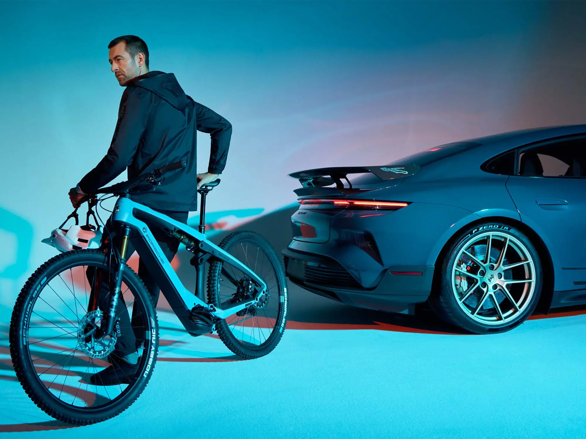 Moški ob Porsche eBike Cross Performance EXC v barvi Provence poleg modrega Porsche Taycana Turbo GT v umetniško osvetljenem prostoru. (David Magazine for Men with Style) / A man with a Porsche eBike Cross Performance EXC in Provence color next to a blue Porsche Taycan Turbo GT in an artistically lit setting. (David Magazine for Men with Style)