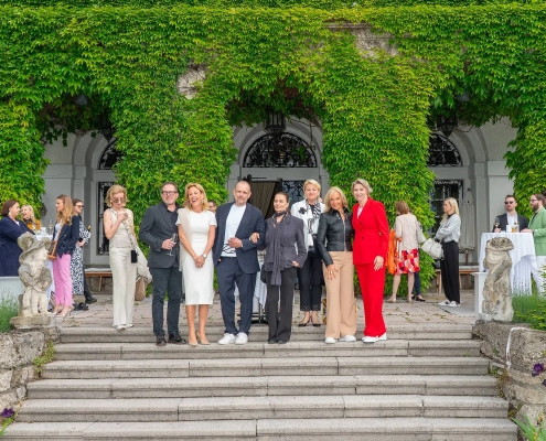 Skupina gostov na stopnišču gradu Leopoldskron na tiskovni konferenci pred prvim amfAR Gala Salzburg 2025; med njimi Gery Keszler, Arabel von Karajan, Elisabeth Gürtler, Marlies Muhr, Marion Pelzl, Elisabeth Friesacher in Mucky Degn. Prispevek revije David – revija za moške s stilom. / Guests gathered on the steps of Schloss Leopoldskron during the press event ahead of the inaugural amfAR Gala Salzburg 2025, including Gery Keszler and other supporters. Featured in David – men's magazine with style.