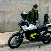 Moški polni električni motocikel Can-Am Origin se polni na ulici – avanturističen e-motor za mestne in terenske vožnje, predstavljen v reviji David za moške s stilom. A man charges the Can-Am Origin electric motorcycle on the street – an adventurous e-bike for urban and off-road riding, featured in David magazine for men with style.