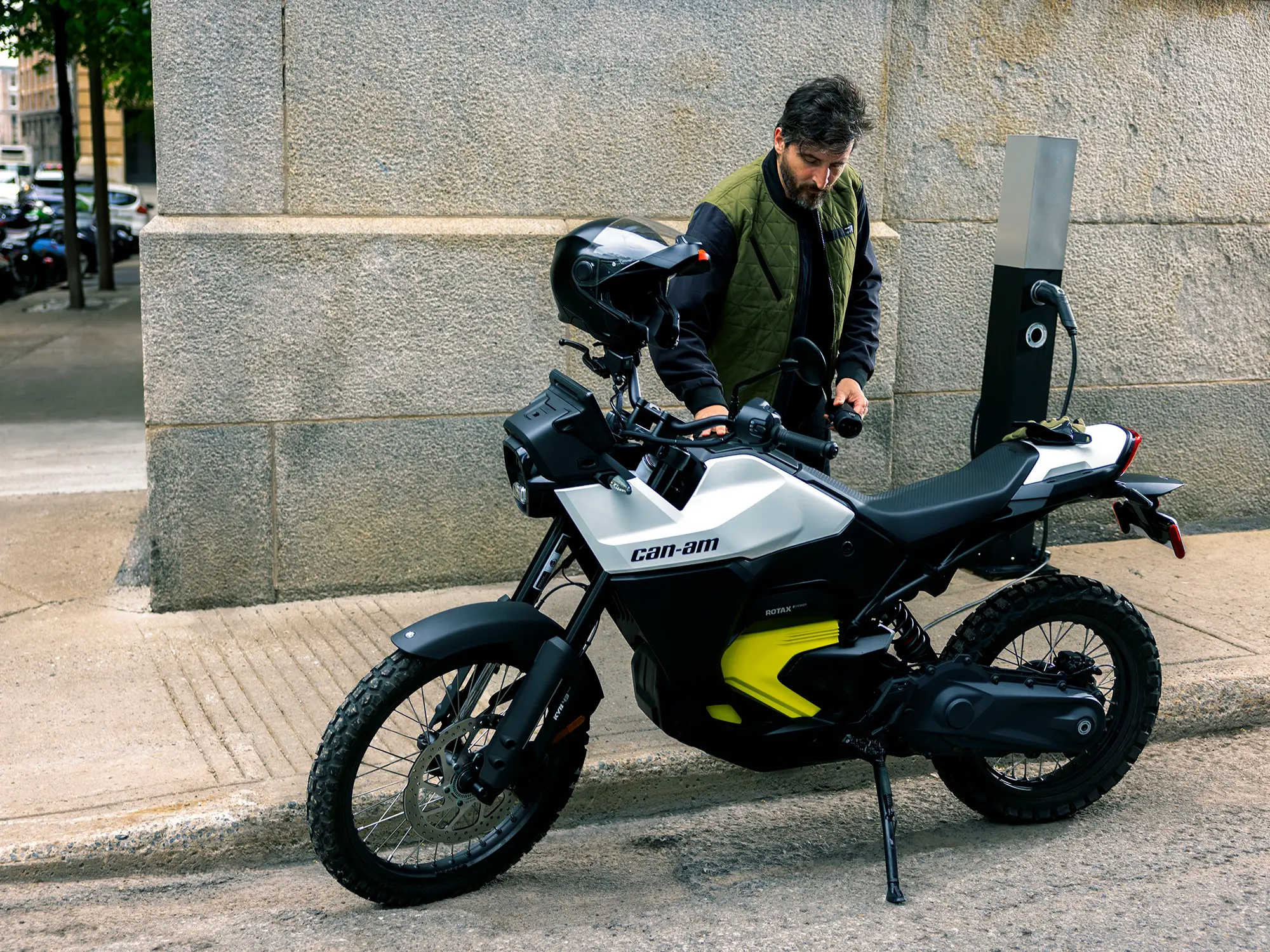 Moški polni električni motocikel Can-Am Origin se polni na ulici – avanturističen e-motor za mestne in terenske vožnje, predstavljen v reviji David za moške s stilom. A man charges the Can-Am Origin electric motorcycle on the street – an adventurous e-bike for urban and off-road riding, featured in David magazine for men with style.