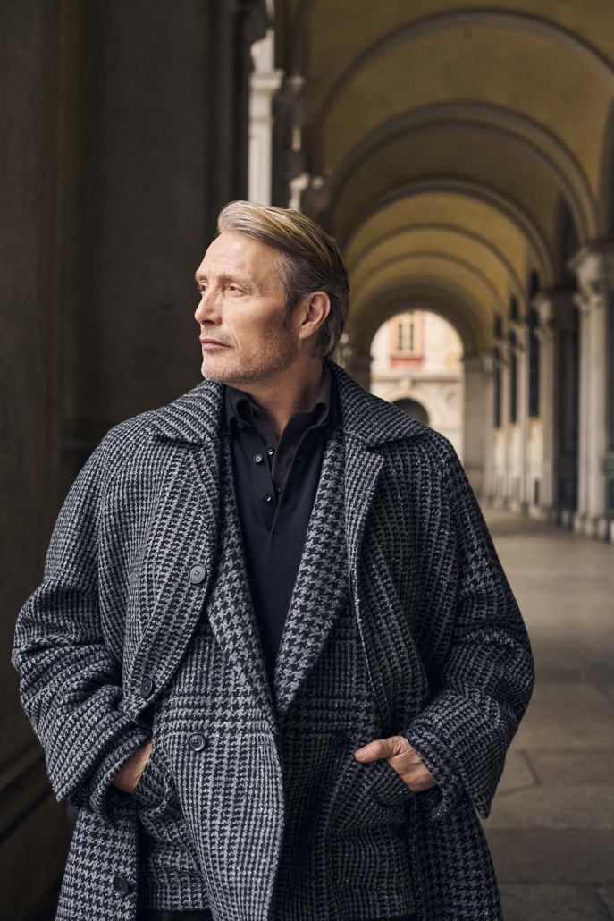 Igralec Mads Mikkelsen oblečen v elegantno pepita vzorčasto ZEGNA obleko, sedi v klasični italijanski kavarni in pije kavo – sofisticirana interpretacija torinskega stila za jesen/zimo 2025.
Actor Mads Mikkelsen wearing an elegant houndstooth ZEGNA suit, seated in a classic Italian café sipping coffee – a sophisticated take on the Turin style for Fall/Winter 2025.