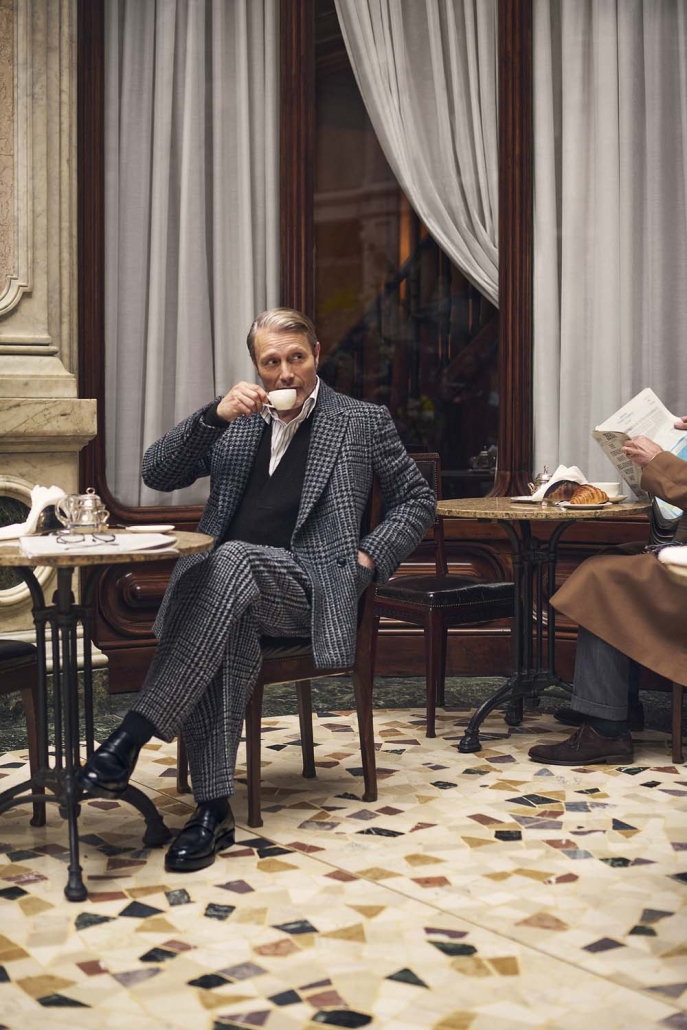 Igralec Mads Mikkelsen oblečen v elegantno pepita vzorčasto ZEGNA obleko, sedi v klasični italijanski kavarni in pije kavo – sofisticirana interpretacija torinskega stila za jesen/zimo 2025.
Actor Mads Mikkelsen wearing an elegant houndstooth ZEGNA suit, seated in a classic Italian café sipping coffee – a sophisticated take on the Turin style for Fall/Winter 2025.