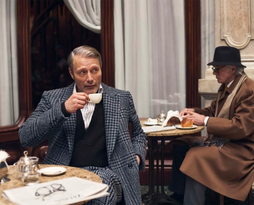 Igralec Mads Mikkelsen oblečen v elegantno pepita vzorčasto ZEGNA obleko, sedi v klasični italijanski kavarni in pije kavo – sofisticirana interpretacija torinskega stila za jesen/zimo 2025. Actor Mads Mikkelsen wearing an elegant houndstooth ZEGNA suit, seated in a classic Italian café sipping coffee – a sophisticated take on the Turin style for Fall/Winter 2025.