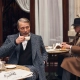 Igralec Mads Mikkelsen oblečen v elegantno pepita vzorčasto ZEGNA obleko, sedi v klasični italijanski kavarni in pije kavo – sofisticirana interpretacija torinskega stila za jesen/zimo 2025. Actor Mads Mikkelsen wearing an elegant houndstooth ZEGNA suit, seated in a classic Italian café sipping coffee – a sophisticated take on the Turin style for Fall/Winter 2025.