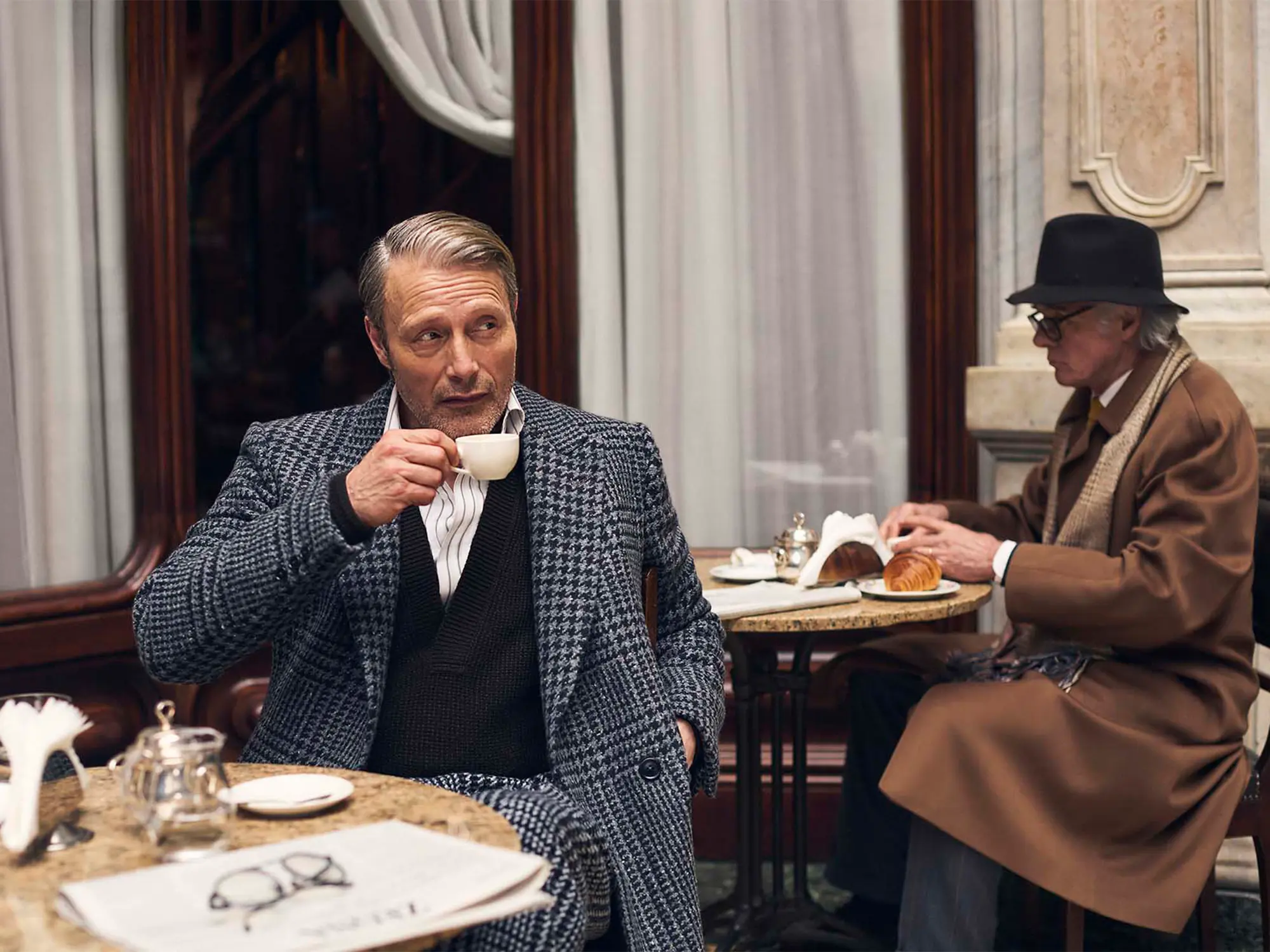 Igralec Mads Mikkelsen oblečen v elegantno pepita vzorčasto ZEGNA obleko, sedi v klasični italijanski kavarni in pije kavo – sofisticirana interpretacija torinskega stila za jesen/zimo 2025. Actor Mads Mikkelsen wearing an elegant houndstooth ZEGNA suit, seated in a classic Italian café sipping coffee – a sophisticated take on the Turin style for Fall/Winter 2025.