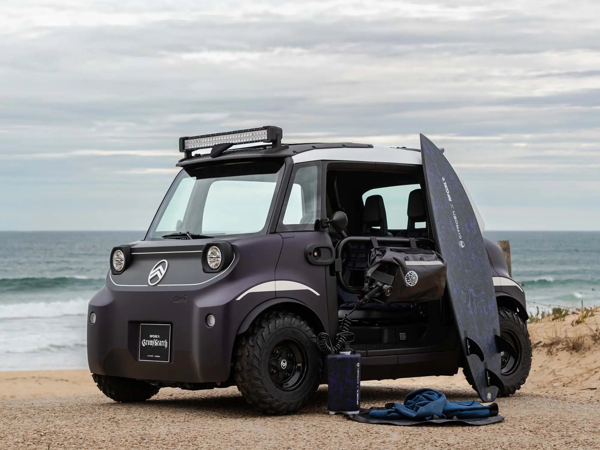 Električni Citroën Ami Buggy Rip Curl parkiran ob obali s surfarsko opremo.