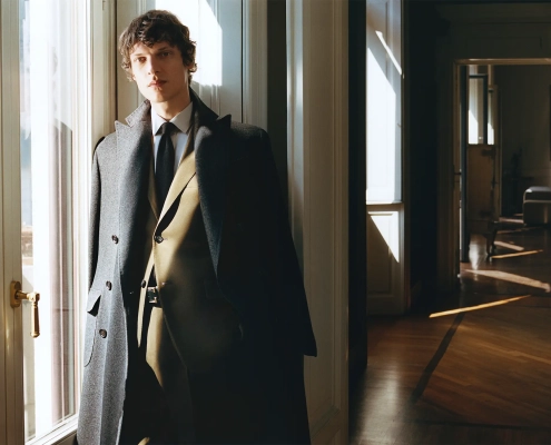 Moški v elegantnem plašču Corneliani stoji ob oknu, oblečen v klasično obleko iz kolekcije jesen–zima 2025, ki odraža italijansko krojenje in prefinjen slog.