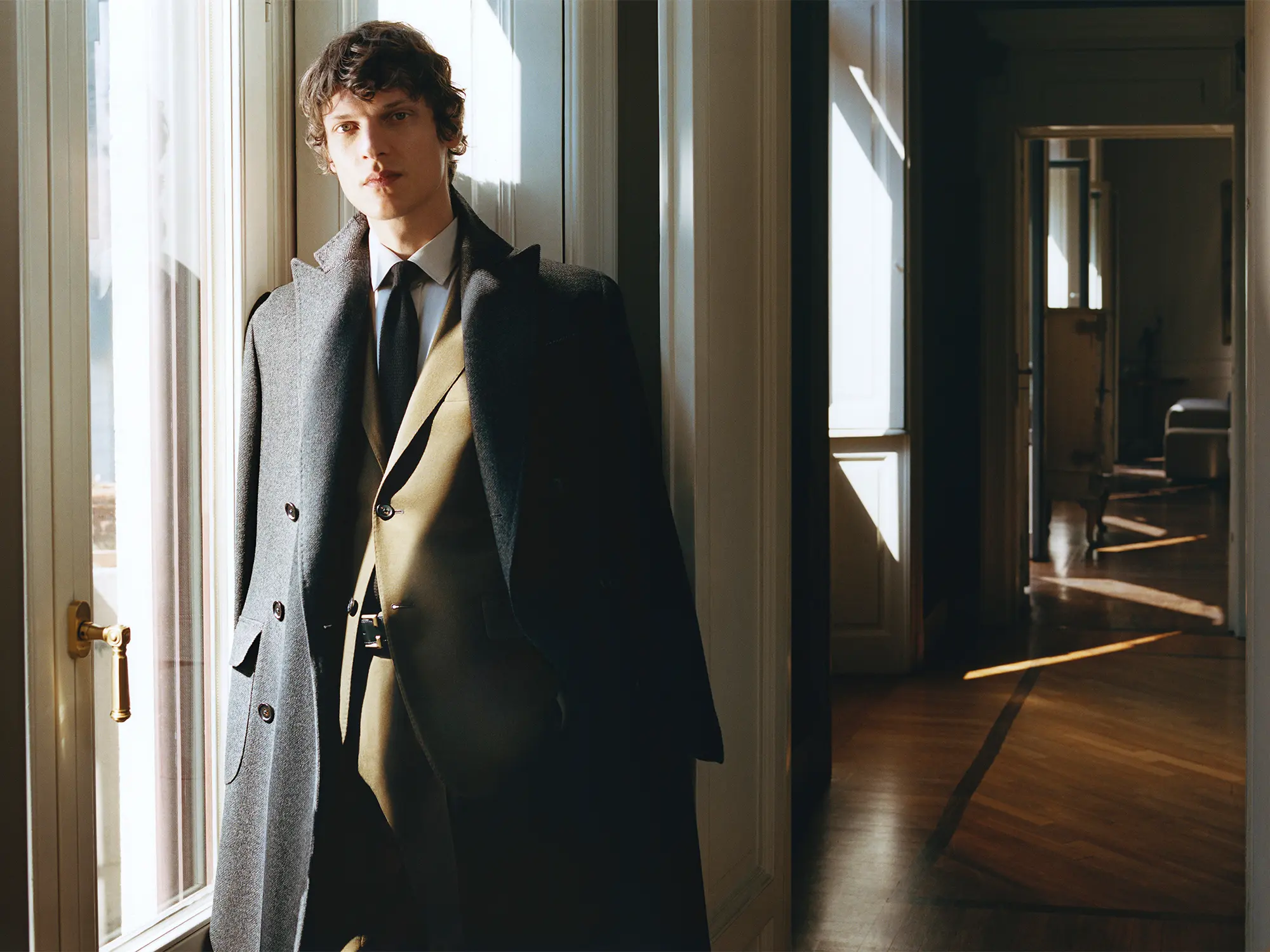 Moški v elegantnem plašču Corneliani stoji ob oknu, oblečen v klasično obleko iz kolekcije jesen–zima 2025, ki odraža italijansko krojenje in prefinjen slog.