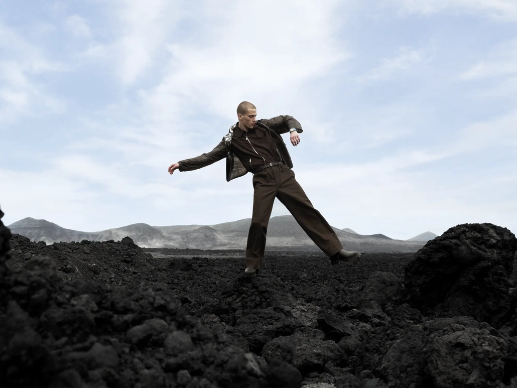Model hodi po temnem skalnatem terenu pod svetlim nebom v kampanji Hermès H08, ki simbolizira odnos med časom, gibanjem in naravo.