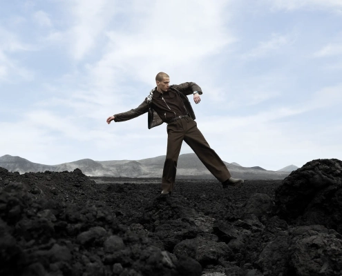 Model hodi po temnem skalnatem terenu pod svetlim nebom v kampanji Hermès H08, ki simbolizira odnos med časom, gibanjem in naravo.