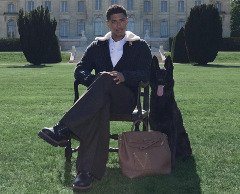 Eleganten moški sedi na stolu na vrtu pred razkošno zgradbo, ob njem črn pes in rjava moška torba Louis Vuitton LV Touch.