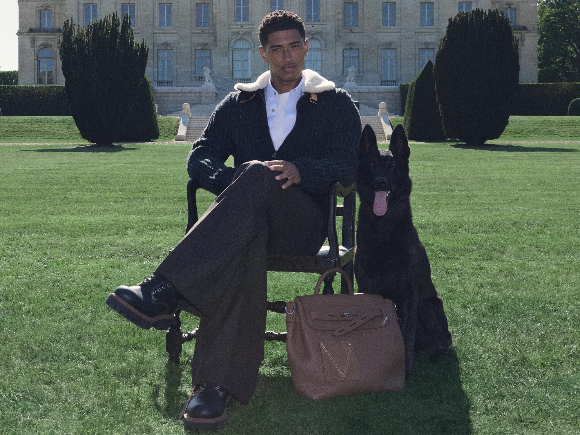 Eleganten moški sedi na stolu na vrtu pred razkošno zgradbo, ob njem črn pes in rjava moška torba Louis Vuitton LV Touch.