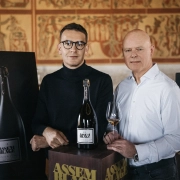 Dve osebi stojita ob veliki steklenici penečega vina Assemblage 211, postavljeni na podstavek, v ozadju pa so vidni dekorativni paneli in predstavitveni materiali znamke.