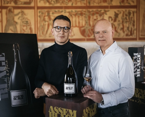 Dve osebi stojita ob veliki steklenici penečega vina Assemblage 211, postavljeni na podstavek, v ozadju pa so vidni dekorativni paneli in predstavitveni materiali znamke.