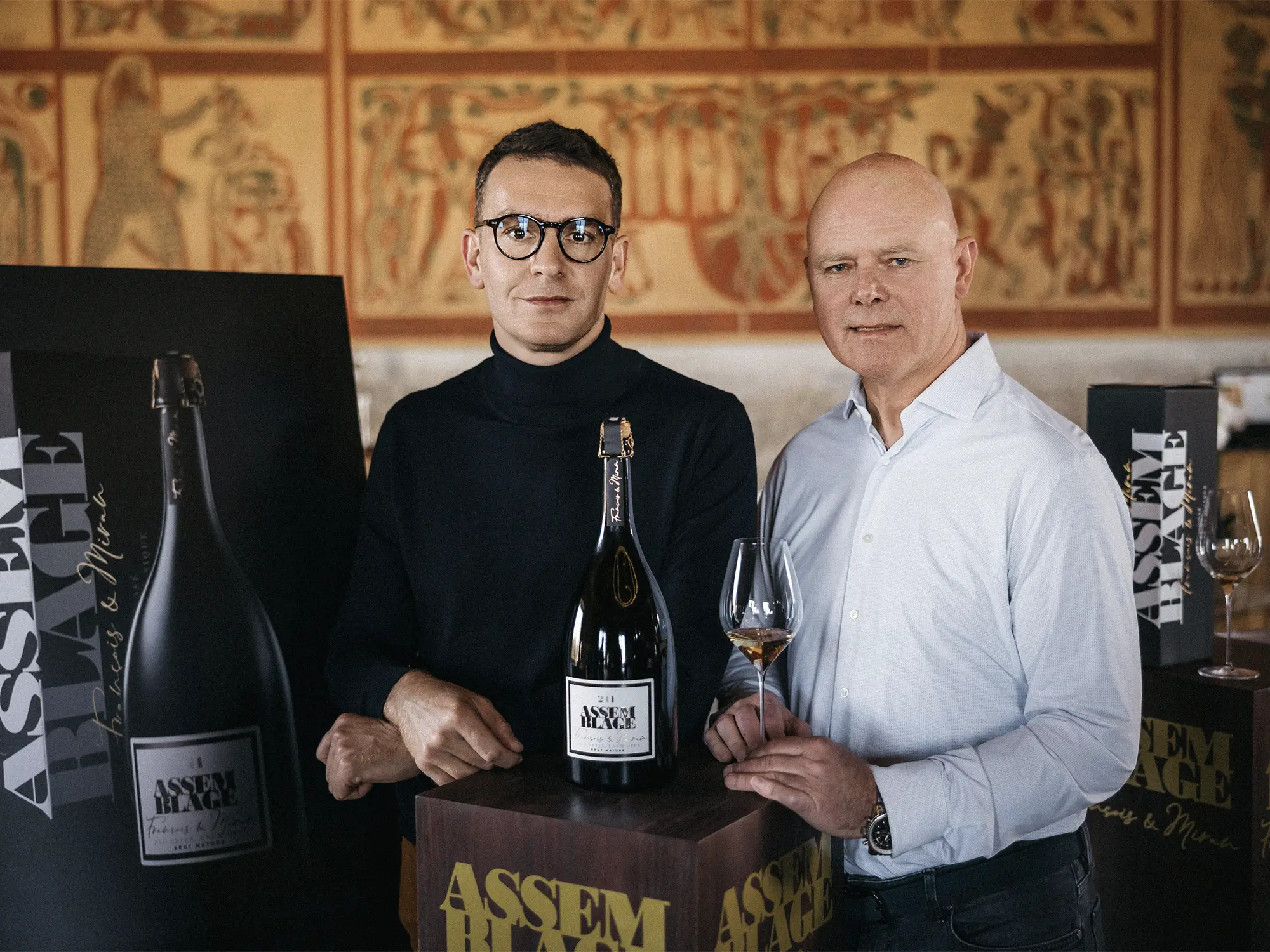 Dve osebi stojita ob veliki steklenici penečega vina Assemblage 211, postavljeni na podstavek, v ozadju pa so vidni dekorativni paneli in predstavitveni materiali znamke.