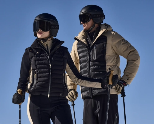 Moški in ženska v smučarskih oblačilih Bogner 007 stojita na snegu s smučarsko opremo.