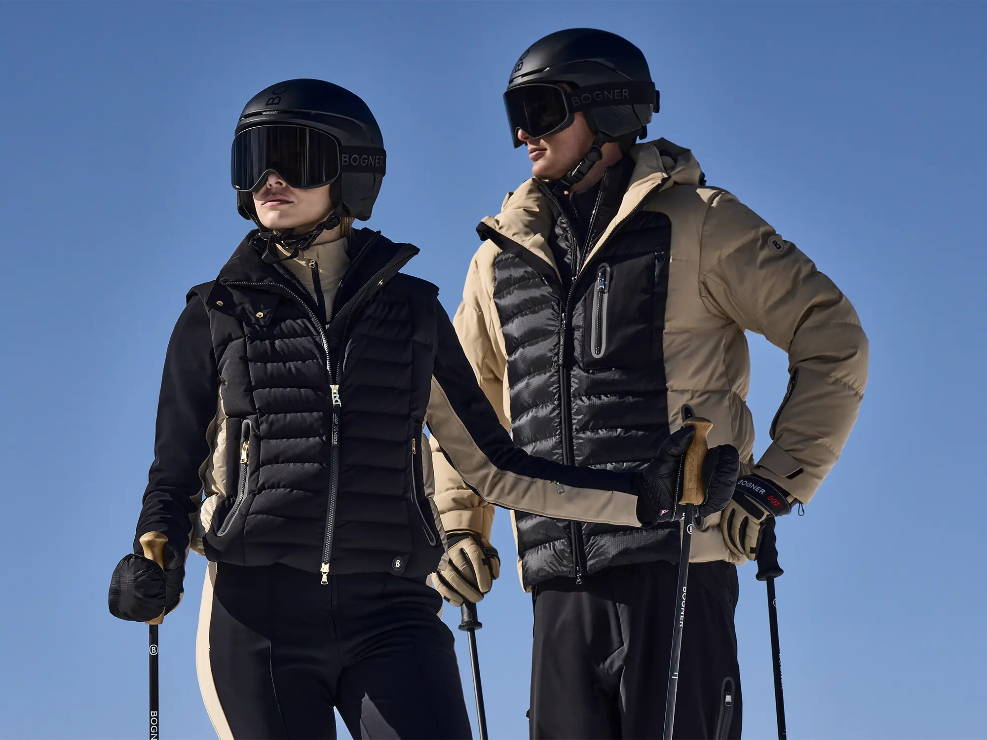 Moški in ženska v smučarskih oblačilih Bogner 007 stojita na snegu s smučarsko opremo.