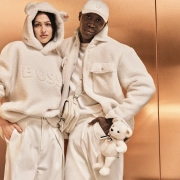 Modela v mehkih kremnih kosih iz kolekcije BOSS x Steiff, oblečena v plišaste jakne in puloverje z medvedkovimi detajli, stojita pred bakreno ozadjem.