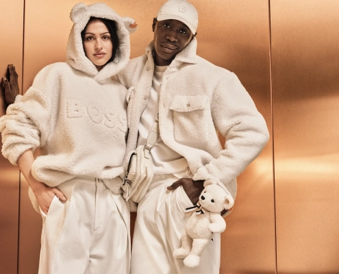 Modela v mehkih kremnih kosih iz kolekcije BOSS x Steiff, oblečena v plišaste jakne in puloverje z medvedkovimi detajli, stojita pred bakreno ozadjem.