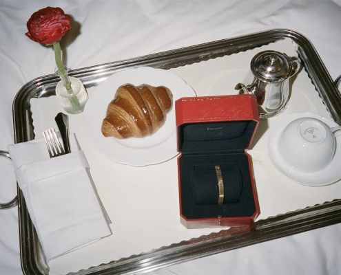 Eleganten zajtrk na srebrnem pladnju z rdečo Cartier škatlico, v kateri je zapestnica Cartier Love, ob kroasanu, skodelici in kovinski posodici za kavo.