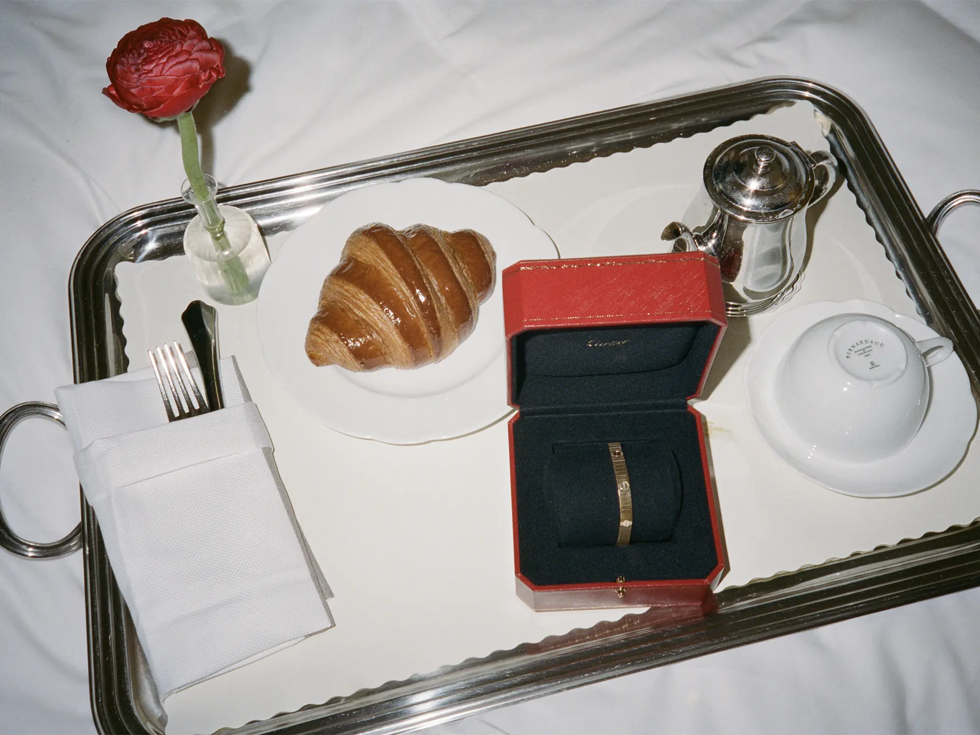 Eleganten zajtrk na srebrnem pladnju z rdečo Cartier škatlico, v kateri je zapestnica Cartier Love, ob kroasanu, skodelici in kovinski posodici za kavo.