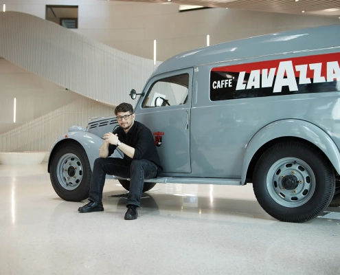 Industrijski oblikovalec Florian Seidl sedi ob klasičnem dostavnem vozilu Lavazza v sodobnem interierju.
