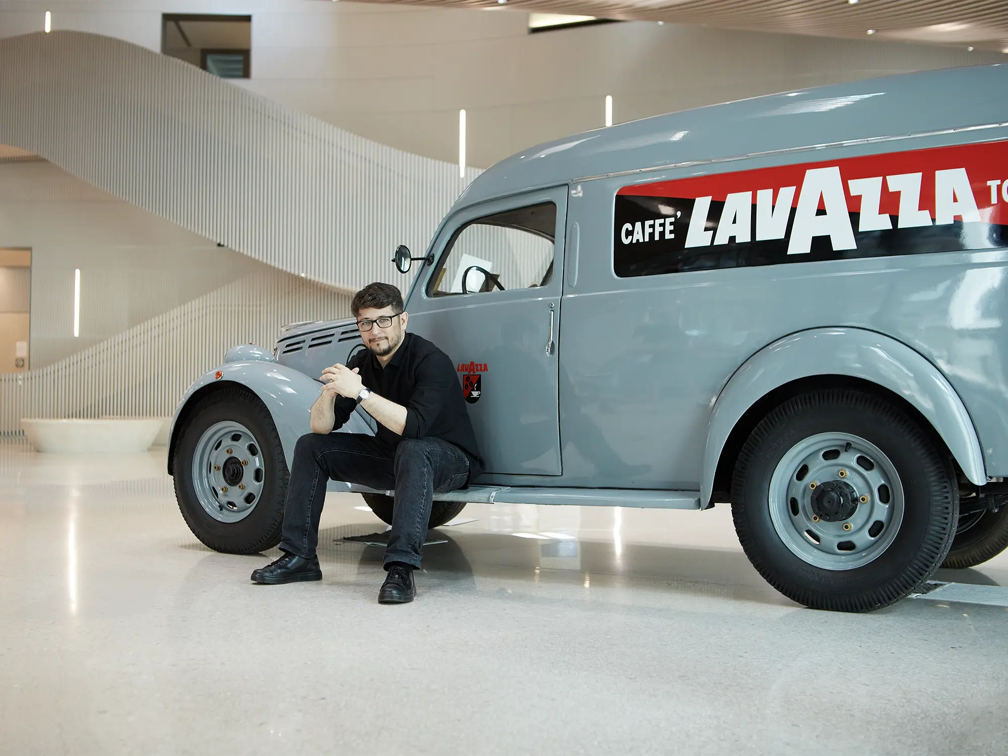 Industrijski oblikovalec Florian Seidl sedi ob klasičnem dostavnem vozilu Lavazza v sodobnem interierju.
