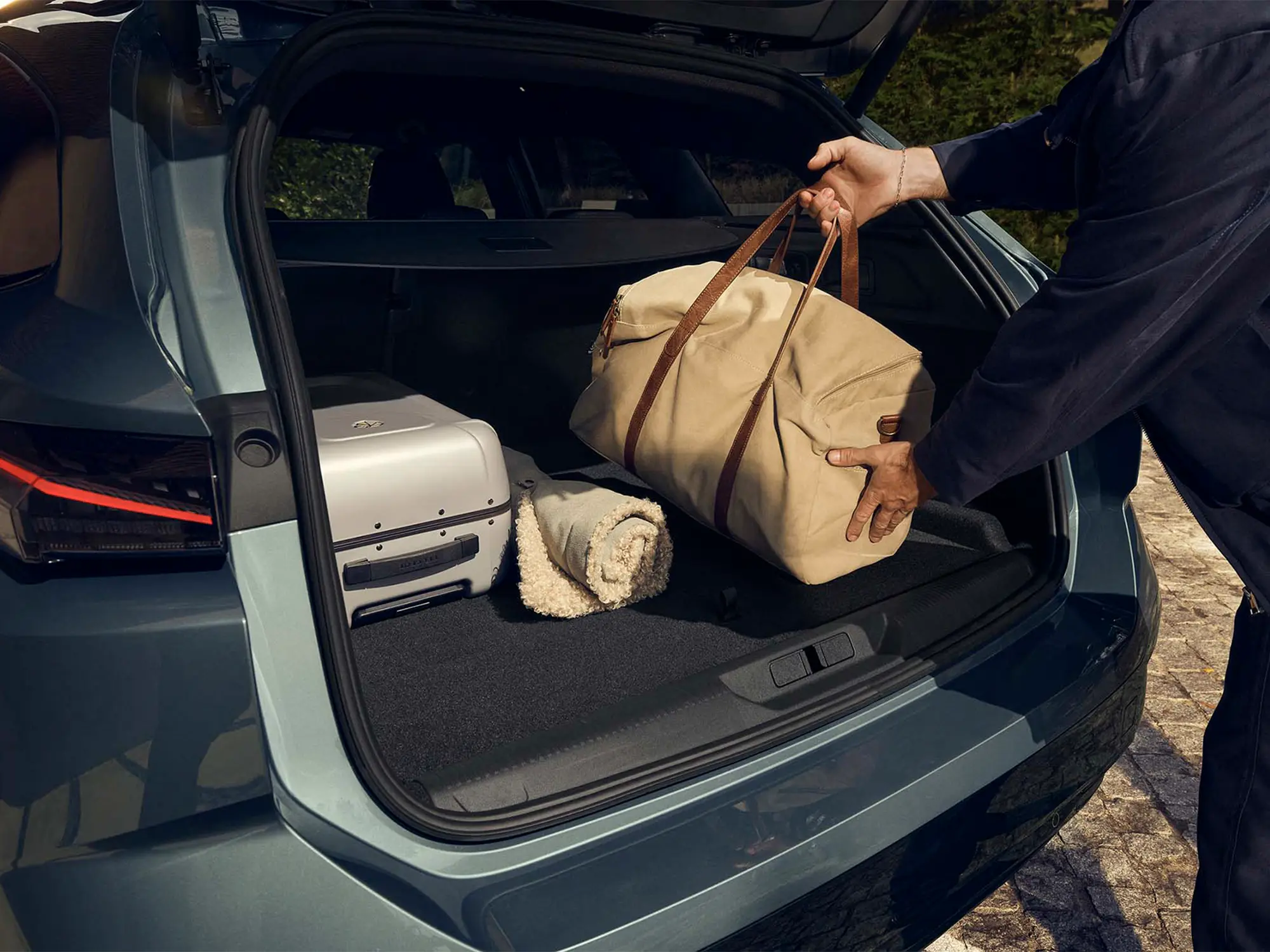 Oseba nalaga potovalno torbo v prostoren prtljažnik novega Peugeota 308 SW, v katerem sta tudi kovček in odeja.